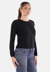 Cardigan nero a maglia con maniche lunghe, scollatura rotonda e chiusura con bottoni. Indossato su jeans blu. Texture liscia e design aderente.