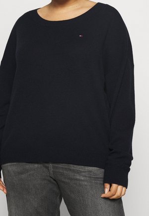 Woman wearing a black long-sleeve sweater with a small red, white, and blue logo on the chest and gray denim jeans.