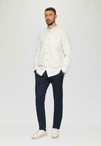 Witte lange mouw knoopsluiting blouse met een zak, donkerblauwe broek en witte sneakers. Eenvoudig ontwerp met een gestructureerde stof en grijze knopen.