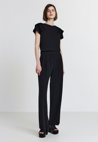 Top nero con maniche arricciate, abbinato a pantaloni neri a vita alta. La modella indossa scarpe platform nere chunky. Tessuto di finitura liscia.