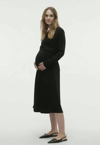 Czarna, długosleeve sukienka ciążowa z dopasowaną górą i zwiewnym dołem, noszona z płaskimi czarnymi butami, na neutralnym tle.