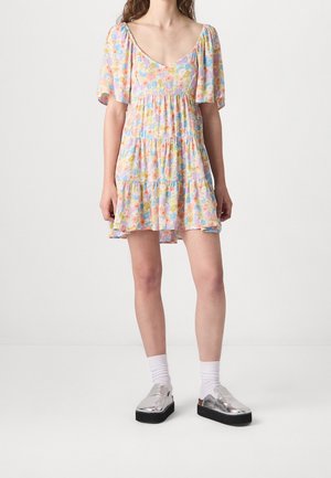 Donna con un vestito corto floreale pastello con maniche a sbuffo, calzini bianchi e scarpe slip-on con plateau argento metallizzato, in piedi su uno sfondo bianco.