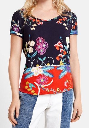 Femme portant un haut à manches courtes bleu marine et rouge avec des motifs floraux colorés, assorti à un jean taille haute bicolore bleu et blanc.