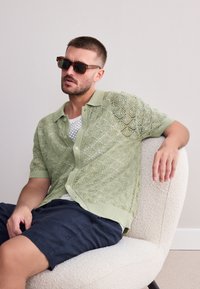 Ljusgrön stickad tröja med korta ärmar och diamantmönster, knäppning fram och ribbad krage, ihop med marinblå shorts. Modell bär solglasögon.