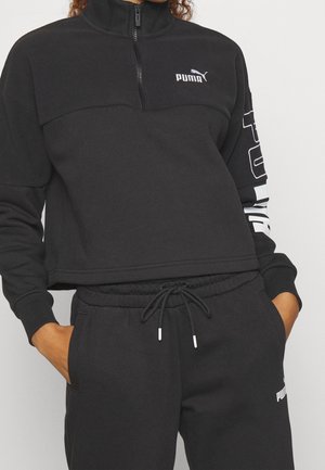 Personne portant un survêtement Puma noir avec un sweat-shirt à demi-fermeture éclair et un pantalon à cordon, mains dans les poches, debout devant un fond uni.