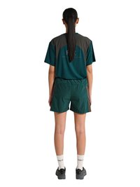 Kvinde med langt mørkt hår i en hestehale iført mørkegrønne og grå sportsshorts og t-shirt, hvide sokker og sorte sportssko, stående.