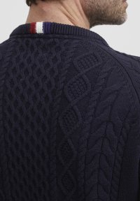 Pull en laine bleu marine avec un design en câble texturé, orné d'une rayure rouge, blanche et violette à l'arrière du col.