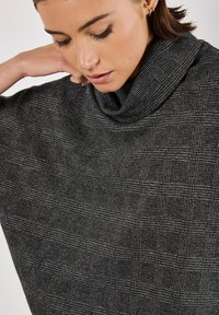 Svart och vit rutigt överdimensionerat poncho med hög krage, som har en texturerad tyg och en draperad silhuett. Ingen synlig hårdvara.