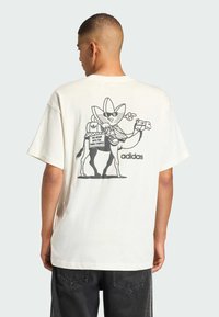 Off-white oversized T-shirt met een zwart cartoonafbeelding van een kameel en een personage met een zonnebril, met het adidas-logo.