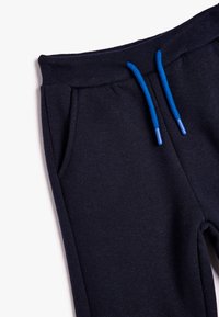 Marineblauwe joggingbroek met een geribbelde tailleband, voorzien van twee zijzakken en blauwe trekkoorden met plastic uiteinden. Soepele stoftextuur.