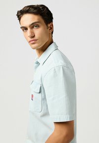  Lichtblauw overhemd met korte mouwen, verticale witte strepen, knoopsluiting, een linkse borstzak en een merklogo label op de zak.