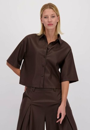 Femme blonde portant une chemise à manches courtes boutonnée brun foncé et un pantalon large assorti, devant un fond clair uni.