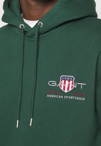 Gros plan d'un sweat à capuche vert foncé avec cordons, logo GANT, emblème en forme de bouclier et texte "1949 American Sportswear" sur la poitrine.