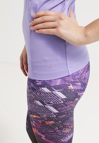 Haut à manches courtes de couleur violet clair avec une texture lisse, associé à des leggings à motifs multicolores présentant des formes géométriques dans des tons foncés.
