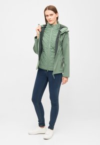 Groene softshell jas met een gewatteerde binnenvoering, hoge kraag, verstelbare capuchon en ritszakken, gedragen over marineblauwe jeans en witte sneakers.