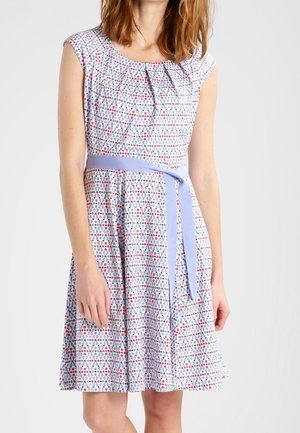 Femme portant une robe sans manches, longueur au genou, avec un motif géométrique en bleu, rouge et blanc, et une ceinture en tissu bleu clair nouée à la taille.