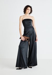 Jumpsuit nero senza spalline con corpetto arricciato, pantaloni a gamba larga e superficie in satin lucido. Abbinato a una pochette metallica e tacchi a punta.