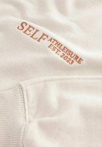 Kreemikas värviga särk, millel on tikkitud tekst "SELF ATHLEISURE EST. 2023" kontrastse heleda pruuniga.