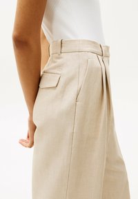Pantaloni beige a pieghe con alta vita, passanti per cintura e tasca posteriore; realizzati in un tessuto leggero e strutturato.