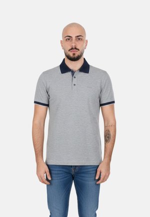 Uomo con la barba che indossa una polo grigia con colletto e bordi delle maniche scuri, fermo su sfondo bianco.