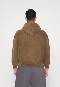 Smeđa hoodie sa teksturiranim završetkom, s kapuljačom na vezanje i rebrastim manžetama, u kombinaciji s sivim hlačama širokog kroja.