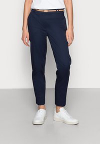 Pantalon tapered bleu marine avec une finition lisse, offrant une coupe slim, des passants de ceinture et un design classique, associé à des baskets blanches.