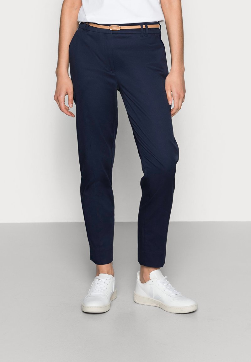 Pantalon tapered bleu marine avec une finition lisse, offrant une coupe slim, des passants de ceinture et un design classique, associé à des baskets blanches.