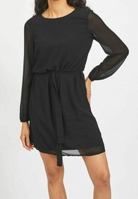 Robe noire avec de longues manches transparentes, un col rond, une taille cintrée avec une ceinture à nouer, et une silhouette fluide.