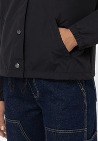 Giacca nera con bottoni a pressione, polsini elasticizzati e coulisse in vita. Indossata sopra pantaloni in denim scuro con cuciture delle tasche visibili.