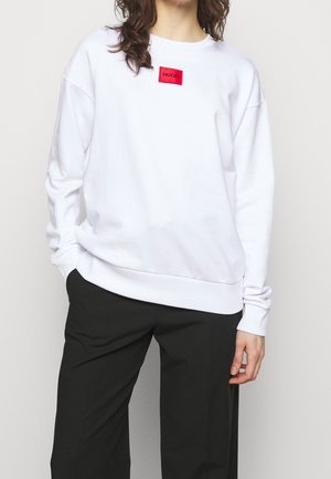 Person iført en løs hvid sweatshirt med en rød "HUGO" logo-patch og sorte bukser, stående med den ene hånd i lommen.