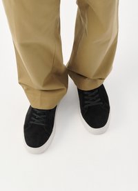 Svarte semskede sneakers med grå snørebånd og hvite gummisåler, kombinert med khaki-bukser som viser en avslappet passform og løse mansjetter.