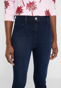 Pantalones de mezclilla azul oscuro con cintura alta, con bolsillos frontales y cierre de botón plateado. Textura suave con un ligero estiramiento.