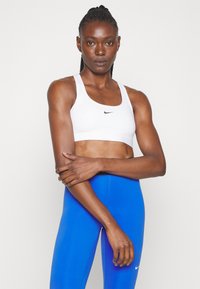 White sports bra with a black Nike logo, paired with blue leggings, featuring a smooth texture and fitted design. Arms crossed in front.