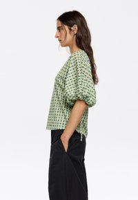 Korte mouwen groene top met opgeblazen mouwen, met een textuurpatroon in verschillende tinten blauw en geel. Draag met donkere broek, zij-aanzicht.