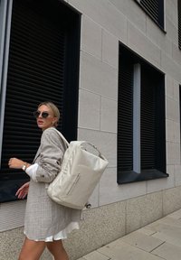 Zaino bianco realizzato in materiale liscio, con una tasca frontale con zip e una maniglia superiore, indossato con un blazer grigio chiaro contro un muro testurizzato.