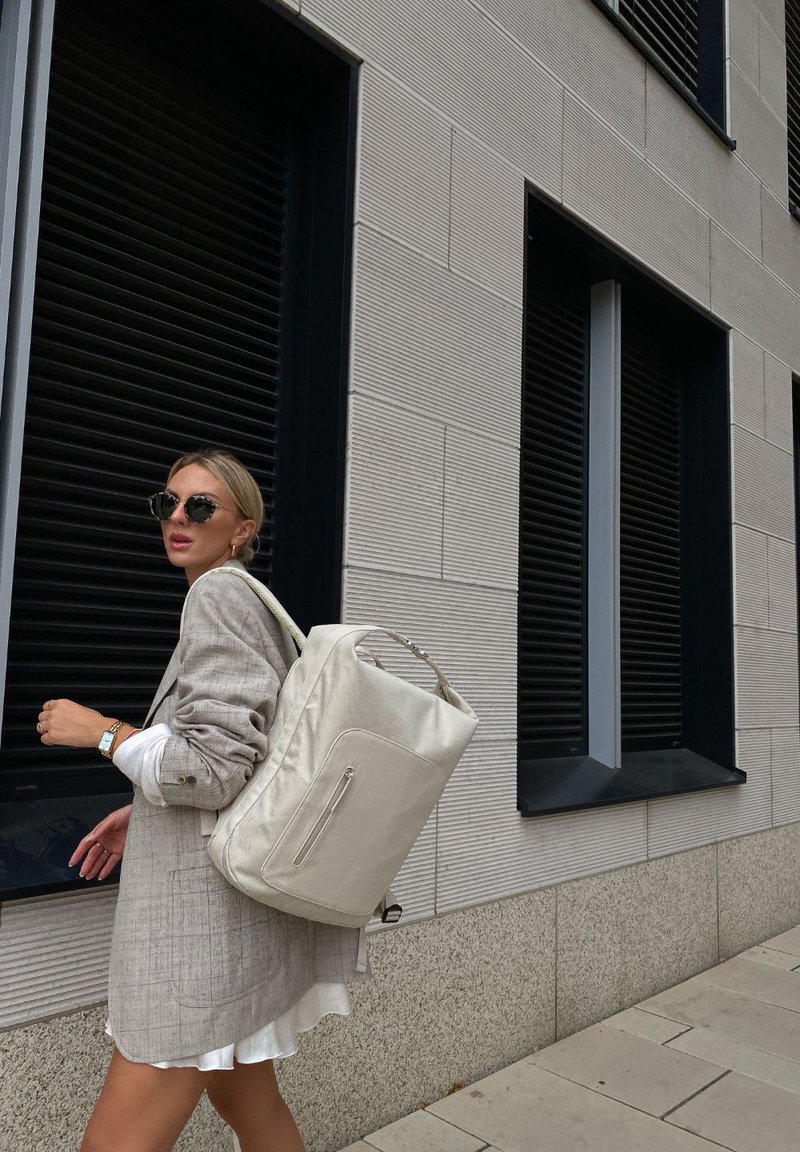 Zaino bianco realizzato in materiale liscio, con una tasca frontale con zip e una maniglia superiore, indossato con un blazer grigio chiaro contro un muro testurizzato.