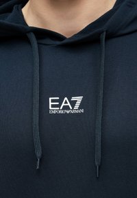 Felpa con cappuccio blu navy con coulisse, materiale morbido e logo bianco EA7 Emporio Armani sul petto. Semplice, con una texture liscia.