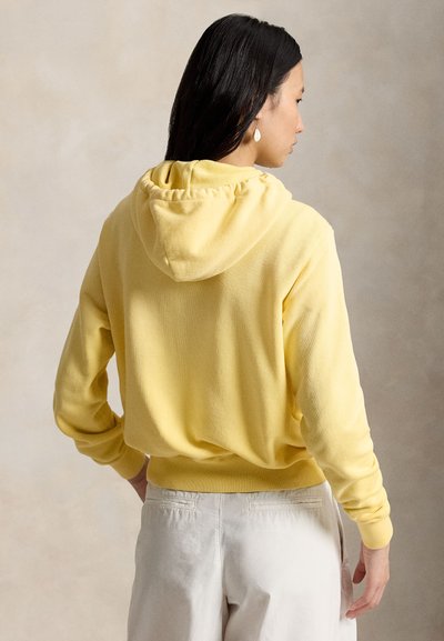 Femme aux longs cheveux foncés portant un sweat à capuche jaune et un pantalon de couleur claire, debout les mains sur les hanches, vue de dos sur un fond neutre.