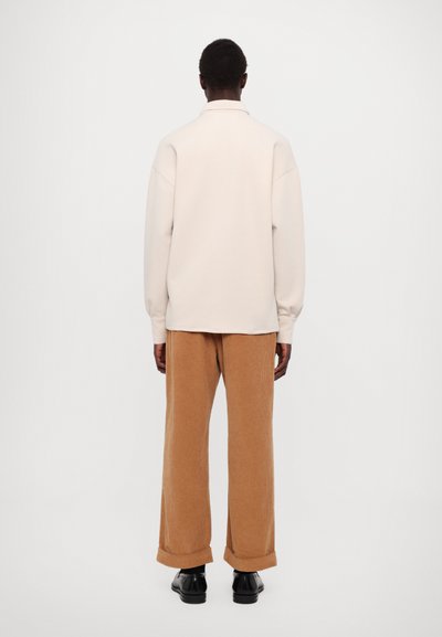 Gai bēša džemperis ar nolīstām plecu šuvēm, saskaņots ar brūniem korduroja biksēm, kurām ir plaša piegriezums un sarullēti piedurknes, papildināts ar melnām kurpēm.