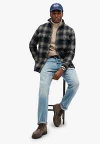 Rutede flannel skjorte i sort og creme, beige strikket sweater, blå jeans, brune støvler og en blå kasket med logo. Står på en skammel.