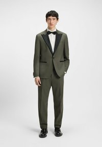 Olivgrüner Anzug mit schwarzem Satin-Revers, ausgestattet mit zwei vorderen Taschen und einer schlanken Passform. Kombiniert mit einem weißen Hemd und einer schwarzen Fliege.