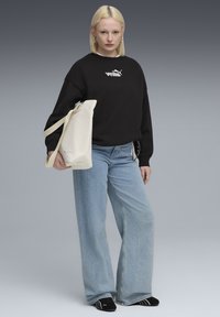 Felpa nera con logo Puma, jeans a gamba larga blu chiaro e calzini neri con strisce bianche. Borsa tote crema tenuta con una mano.