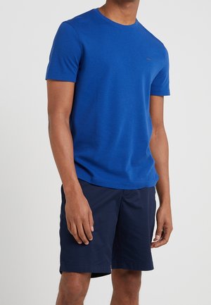 Un homme portant un t-shirt uni bleu royal à manches courtes et un short bleu marine jusqu'aux genoux, sur un fond clair.