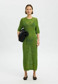 Vestido verde de punto con mangas tres cuartos, que presenta un patrón geométrico y un acabado texturizado. La modelo sostiene un bolso negro texturizado.