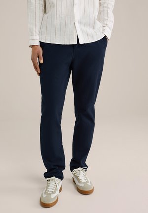 Pantalon texturé bleu marine avec une coupe décontractée ; associé à des baskets gris clair et blanches avec des semelles marron. Chemise à rayures blanches visible.