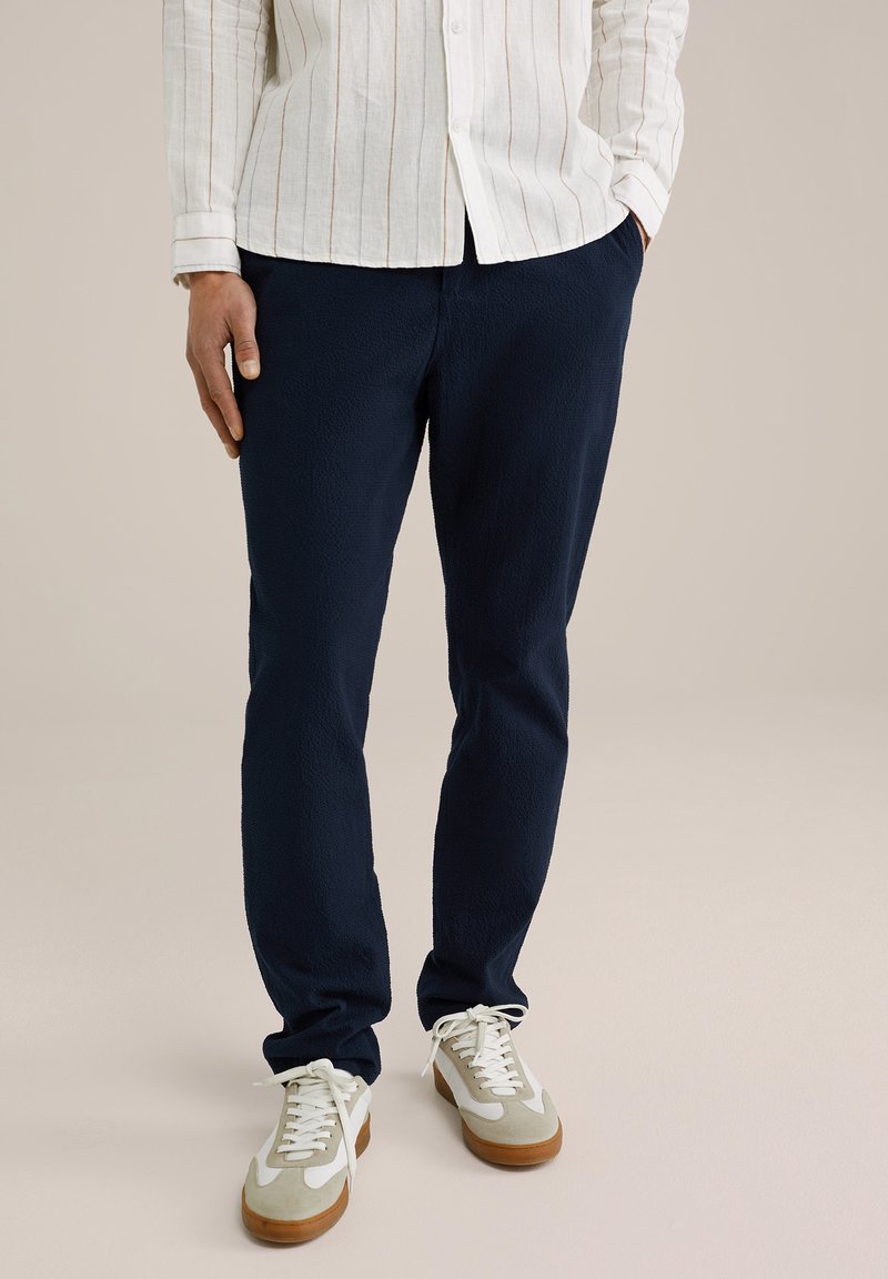 Jūras zilas teksturētas bikses ar brīvu piegriezumu; savienotas ar gaiši pelēkiem un baltajiem sporta apaviem ar brūniem zoliem. Redzama balta svītrainā krekls.