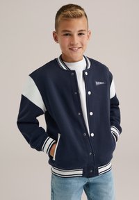 Mörkblå varsityjacka med vita ärmar, ribbade mudd och krage. Har tryckknappar och en liten logopatch på bröstet.