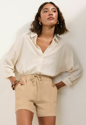 Kvinde med skulderlangt hår iført en løs cremefarvet skjorte med knapper, beige shorts med snøre, guld hoop øreringe og armbånd, stående med hænderne i lommerne.