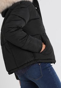 Svart vadderad jacka med fuskpälskrage, quiltad textur, sidofickor och figurnära passform, kombinerad med mörkblå jeans.