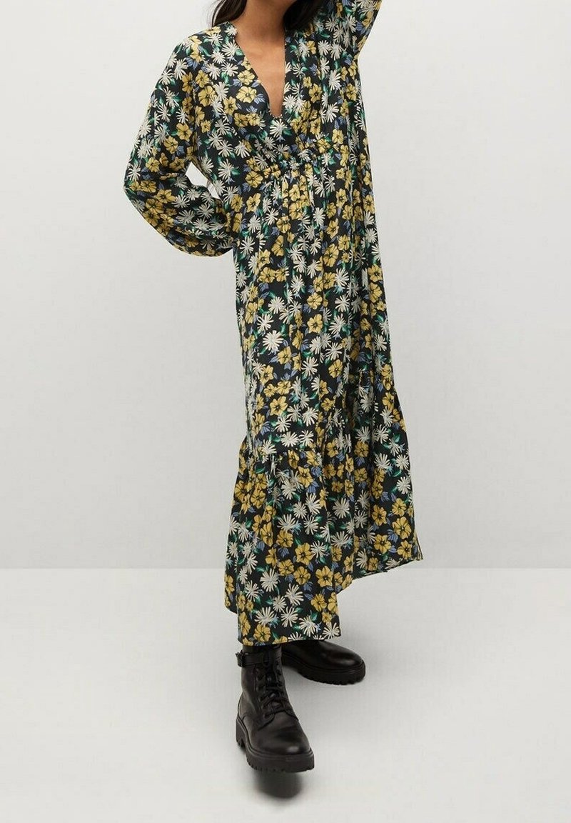Femme portant une longue robe fleurie avec des fleurs jaunes et blanches et des bottes noires à lacets, posant devant un fond uni.
